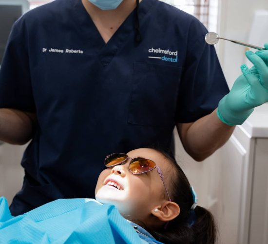 child laying in dentist chair with dentist glasses on while dentist checks her teeth